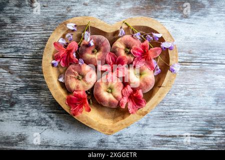 Pfirsiche in einer herzförmigen Holzschale auf einem Tisch mit verstreuten Sommerblumen Stockfoto