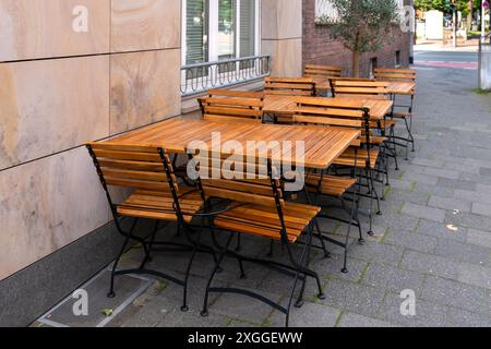Eine Reihe von Holztischen und -Stühlen steht auf einer Kopfsteinpflasterstraße vor einem Restaurant oder Café. Die Tische sind mit leeren Stühlen gedeckt, um Gäste willkommen zu heißen. Das warme Sonnenlicht und die charmante europäische Architektur schaffen eine herrliche Atmosphäre. Stockfoto