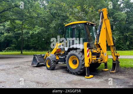 Ein gelber Baggerlader wird auf einem Schotterweg in einer Waldlage geparkt. Der Baggerladerarm wird angehoben und die Schaufel liegt auf dem Boden auf. Stockfoto