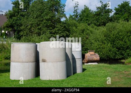 Betonzylinder sitzen auf einem grasbewachsenen Feld in der Nähe einer Baumgruppe. Im Hintergrund befindet sich ein rostiger Baggerlöffel. Stockfoto