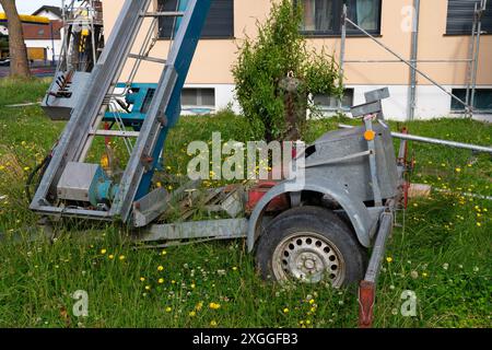 Ein Bauanhänger mit blauem Elektrolift parkt auf einem grasbewachsenen Gelände in der Nähe eines Gebäudes mit Gerüsten. Stockfoto