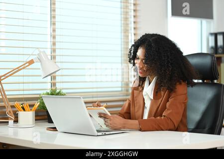 Lächelnde afroamerikanische Geschäftsfrau in brauner Jacke, die mit dem Laptop arbeitet und am Schreibtisch Notizen macht Stockfoto