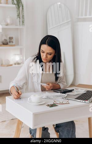 Selbstbewusste junge Frau, die am Schreibtisch mit Laptop, Dokumenten und Taschenrechner arbeitet. Schreiben von Notizen bei der Prüfung von Papieren. Verwaltung von Zahlungen und Steuern für Versorgungsunternehmen Stockfoto