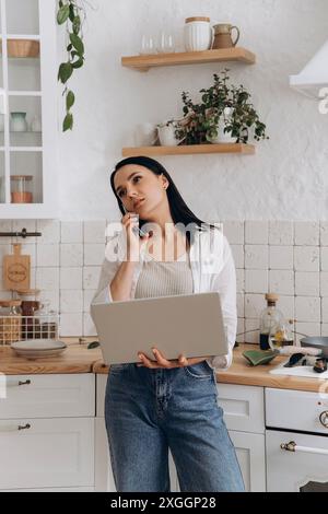Irritierte junge Frau beim Multitasking, arbeitet widerwillig am Laptop und telefoniert an einem Wochenende im Kithen. Konzept der Diskussionen über Stockfoto