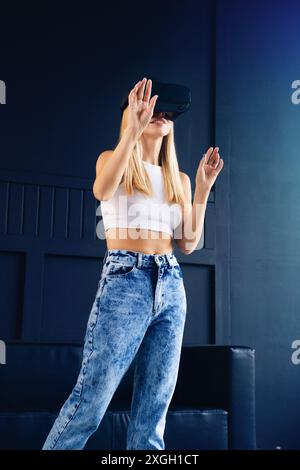 Die junge blonde Frau in weißem Crop Top verwendet ein Virtual-Reality-Headset in einem Studio und streckt mit den Händen aus, um mit der Umgebung zu interagieren Stockfoto