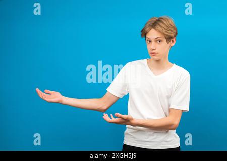 Der junge Teenager zeigt die Handflächen isoliert auf blau zur Seite. Freundlicher, gut aussehender Junge, der seine Aufmerksamkeit auf den Kopierbereich, das beste Angebot, den Verkauf und die Präsentation von Neuheit lenkt Stockfoto