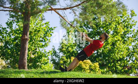 Muskulöser Mann beim TRX-Workout im üppigen grünen Park, der Baumäste zur Unterstützung verwendet und perfekte Form und Konzentration demonstriert. Stockfoto