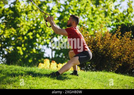 Sportlicher Mann mit gesungenem muskulösen Körper, in Sportbekleidung trainierende Beine mit trx Fitnessgurten im Freien gegen grünen Park Stockfoto