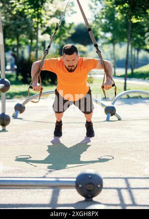 Bärtiger Mann in orangefarbenem T-Shirt und schwarzen Shorts beim trx-Training mit Trageriemen im Park. Liegestütze Stockfoto