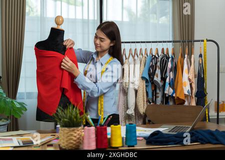 Glückliche, lächelnde asiatische kreative Modedesignerin arbeitet in ihrer Schneiderei. Frau, die ein neues Kleidungskonzept entwickelt. Stockfoto