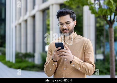 Junger Geschäftsmann, der außerhalb des Bürogebäudes ein Smartphone nutzt und sich auf das Gerät konzentriert. Legere Kleidung, modernes Unternehmensumfeld, professioneller Lebensstil. Städtisches Äußere, verbunden Stockfoto