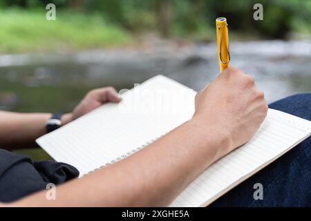Ein junger Mann macht Notizen in einem Notizbuch über die Natur. Der Mann sitzt an einem Baum mit dem Rücken zu ihm, ein Bleistift und ein Notizbuch in der Hand, macht Notizen Stockfoto