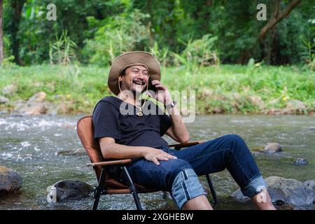 Bärtiger junger Mann, der eine Pause macht, sein Smartphone anmacht und auf einem Handy spricht, der auf einem Stuhl liegt, mitten in einem sonnigen Fluss draußen Stockfoto