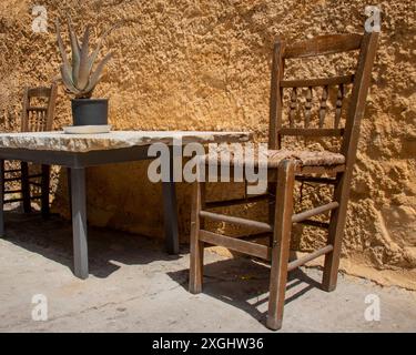 Alter, klappriger Holzstuhl im Freien neben einem Steintisch mit einer Kaktuspflanze und einer strukturierten Terrakotta-Wand im Sonnenschein Griechenlands Stockfoto