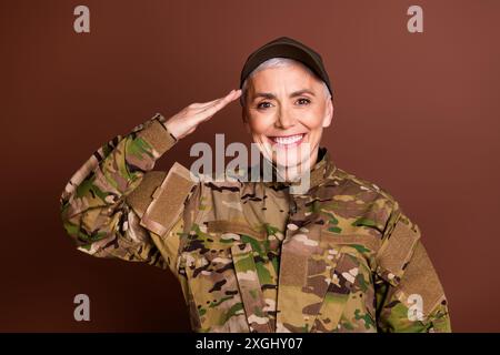 Foto einer attraktiven lächelnden älteren Dame in Camo-Uniform, die Ihnen Ehre Geste leerer Raum isolierter brauner Hintergrund zeigt Stockfoto