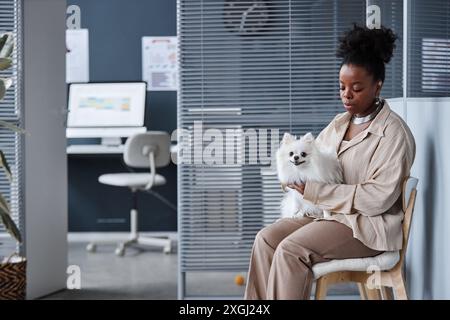 Weitwinkelaufnahme einer lockigen Afroamerikanerin mit weißem pommerschen Hund an Händen, die im Wartebereich der Tierarztklinik sitzt, Kopierraum Stockfoto