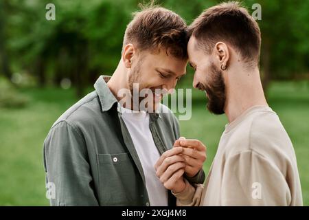 Zwei bärtige schwule Männer stehen in einem grünen Park dicht beieinander, ihre Köpfe berühren sich, während sie einen zärtlichen Moment teilen. Stockfoto