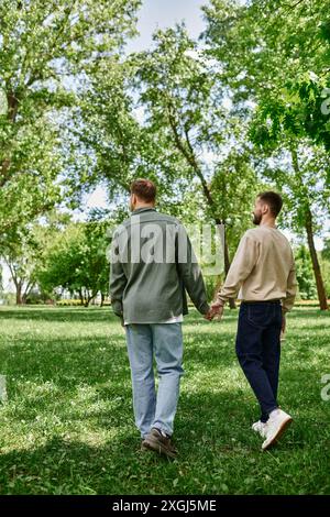 Ein schwules Paar mit Bärten in legeren Kleidern geht Hand in Hand durch einen grünen Park. Stockfoto