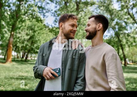 Zwei bärtige Männer in lässiger Kleidung stehen in einem grünen Park dicht beieinander und teilen einen liebevollen Moment. Stockfoto