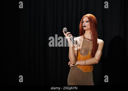 Ein Drag-Performer mit langen roten Haaren und einem glamourösen Outfit hält ein Mikrofon vor einem schwarzen Vorhang. Stockfoto