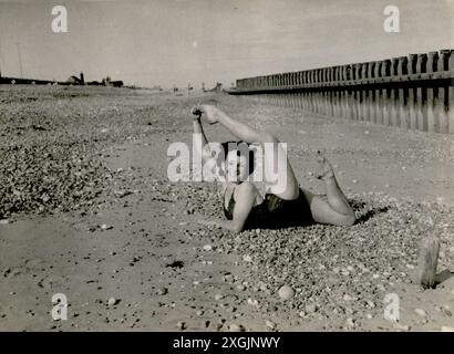 1940er Jahre, historisch, eine junge Frau im Badeanzug, die auf ihrem Bauch an einem steinigen Strand lag, ihr linkes Bein über dem Kopf hielt, ihre Zehen mit der rechten Hand hielt, was ihre Flexibilität und ihren geschmeidigen Körper zeigte. Stockfoto