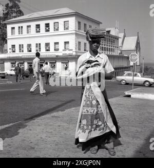 1950er Jahre, historisch, draußen auf der Straße, ein junger einheimischer Mann, der lokale Textilien, Tischdecken verkauft, seinen Vorrat auf dem Kopf balanciert, Colombo, Ceylon (Sri Lanka). Hinter ihm befindet sich ein Gebäude mit dem Bücherladen der SL Diözese im Erdgeschoss. Stockfoto
