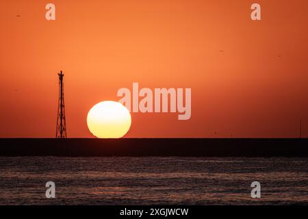 Eine atemberaubende Sonnenaufgangsszene am Strand, mit einem Pier, dem Sonnenaufgang über einer großen Kugel, einem orangen Himmel, Silhouettenvögeln und einer Metallstruktur Stockfoto