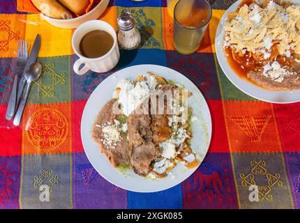 Authentisches mexikanisches Frühstück mit Chilaquiles mit zartem Fleisch, frischen Bohnen und frischem Käse, serviert mit einer Tasse dampfendem Kaffee. Vibra Stockfoto