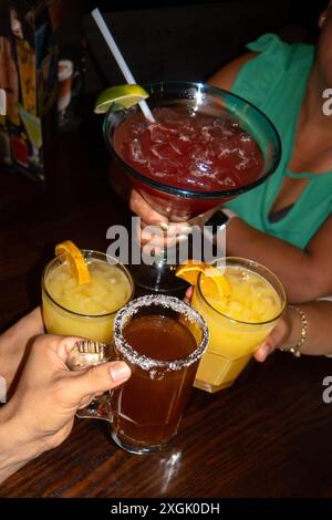 Lebhaftes Bild von Freunden, die mit einer Vielzahl von Cocktails, darunter eine große Margarita, zwei Orangengetränke und ein Bier mit gesalzenem Rand, toasten. Celebrato Stockfoto