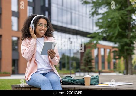 Lächelnde junge Frau, die ein digitales Tablet und Kopfhörer benutzt, während sie draußen auf dem Universitätsgelände sitzt. Genießen Sie Musik und Technik, tragen Sie Freizeitkleidung. Entspannte und fröhliche Atmosphäre an sonnigen Tagen. Stockfoto