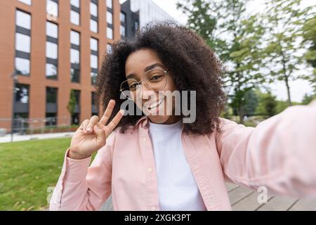 Eine junge Frau mit lockigen Haaren, die Selfie macht und draußen ein Friedenszeichen in der Nähe eines modernen Gebäudes zeigt. Lässige Kleidung, fröhlicher Ausdruck, urbane Umgebung, sonniger Tag. Stockfoto