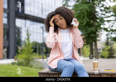 Junge Frau mit lockigem Haar und Brille, die Taschentücher hält und Kopfschmerzen hat. Vor einem modernen Gebäude sitzen und gestresst aussehen. Krankheitsbegriff, Stress und urbaner Lebensstil. Stockfoto