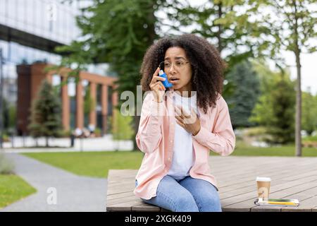 Junge Frau mit Asthma, die Inhalatoren zur Erleichterung benutzt, während sie auf der Bank im Park sitzt. Sie sieht ängstlich aus und trägt legere Kleidung. Grüne Bäume und moderne Gebäude im Hintergrund. Stockfoto