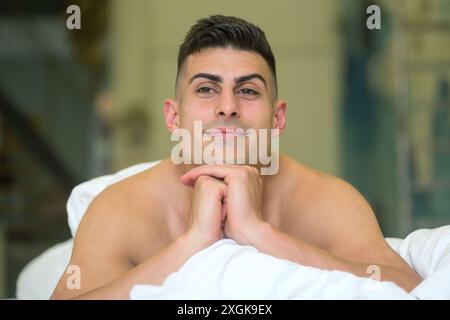 Ein junger Mann mit kurzen dunklen Haaren liegt auf dem Bett, das Kinn auf den Händen liegend, und lächelt zufrieden. Der Hintergrund mit weichem Fokus sorgt für eine ruhige, intime Atmosphäre Stockfoto