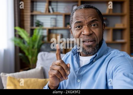 Mann mittleren Alters, der während eines Videoanrufs zu Hause mit dem Finger gestickt hat. Ansprechende und kommunikative Interaktion, die eine Erklärung oder einen Lernmoment suggeriert. Komfortabler Wohnbereich Stockfoto