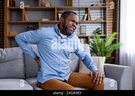 Mann mittleren Alters, der Rückenschmerzen erlebt, während er auf einer bequemen Couch im modernen Wohnzimmer sitzt. Ein Mann, der vor Unbehagen grimmt, sich zurückhält und Erleichterung braucht. Gemütliche, gut beleuchtete Inneneinrichtung. Stockfoto