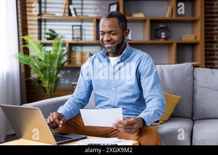 Ein lächelnder Mann, der die Arbeit von zu Hause aus genießt, während er sich Finanzdokumente ansieht. Unternehmer verwalten Geschäftsfinanzen mit Laptop zu Hause in gemütlicher Wohnzimmerumgebung Stockfoto