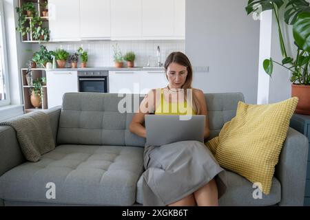 Ernsthaft fokussierte Frau setzt auf dem Sofa auf Laptop und konzentriert sich auf Fernarbeit, um geschäftliche Probleme zu lösen. Stockfoto