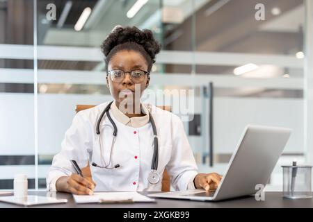 Selbstbewusste Ärztin am Schreibtisch in der modernen Klinik. Der Arzt verwendet Laptop und schreibt Notizen, zeigt Professionalität und Hingabe für die medizinische Praxis. Stockfoto