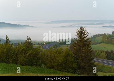 Eine gewundene Autobahn schlängelt sich durch ein in Nebel gehülltes Tal mit üppigen grünen Hügeln und Bäumen im Vordergrund. Stockfoto