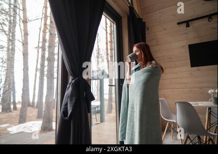 Eine kaukasische Frau, in eine Decke gewickelt, trinkt Kaffee in der Nähe des Terrassenfensters. Stockfoto