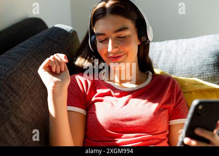 Das kaukasische Teenager-Mädchen genießt zu Hause Musik auf ihren hübschen Kopfhörern. Sie entspannt sich auf einer Couch und nimmt sich im warmen Sonnenlicht mit einem Smartphone in der Hand. Stockfoto