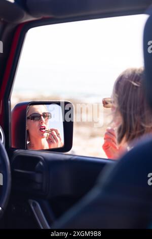 Die junge kaukasische Frau trägt Lippenstift im Autospiegel auf einer Autofahrt, mit Kopierraum. Die Momente sind eine Mischung aus Schönheitsroutine und Reisen. Stockfoto