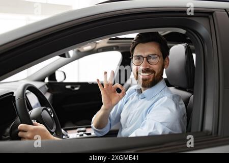 Lächelnder Geschäftsmann sitzt im Auto im Händlerbetrieb und zeigt eine gute Geste Stockfoto