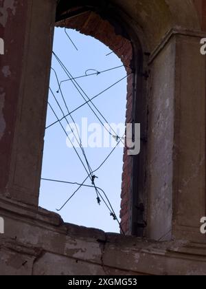 Blick durch ein kaputtes Fenster in einem alten verlassenen Gebäude mit bröckelnden Wänden und freiliegender Ziegelstruktur, gesehen vor einem blauen Himmel. Stockfoto
