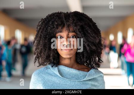 Porträt eines selbstbewussten jungen afroamerikanischen Studenten mit natürlichen lockigen Haaren, der auf einem geschäftigen Universitätscampus steht. Sie trägt einen hellblauen Schweiß Stockfoto