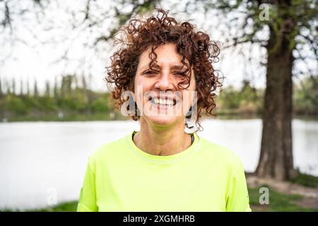 Porträt einer fröhlichen Frau mittleren Alters mit lockigem Haar, die ein hellgelbes Sporthemd trägt und draußen am See steht. Konzept des aktiven Lebensstils Stockfoto