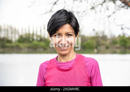 Porträt einer lächelnden Frau mittleren Alters mit kurzen schwarzen Haaren in rosa Sportkleidung, die draußen am See steht. Konzept des aktiven Lebensstils und Stockfoto