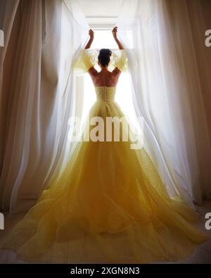 Die Silhouette einer Frau in einem goldenen, luxuriösen Kleid vor dem Hintergrund eines Fensters hält einen Vorhang mit den Händen. Elegante Dame in gelber Seide Stockfoto