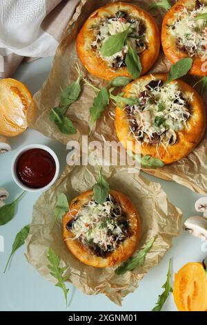 Mehrere Mini-Pizzen mit Käse und Pilzen werden auf Pergamentpapier mit Rucolablättern und Tomatenhälften auf einem hellblauen Tisch serviert Stockfoto